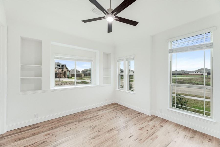 Empty room with hardwood floors, ceiling fan, built-in shelves, and large windows with neighborhood views