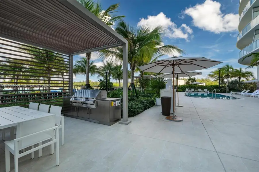 Outdoor kitchen featuring a built-in stainless steel grill and stone-finish countertop beneath a louvered pergola, complemented by large-format tile flooring and an inground swimming pool
