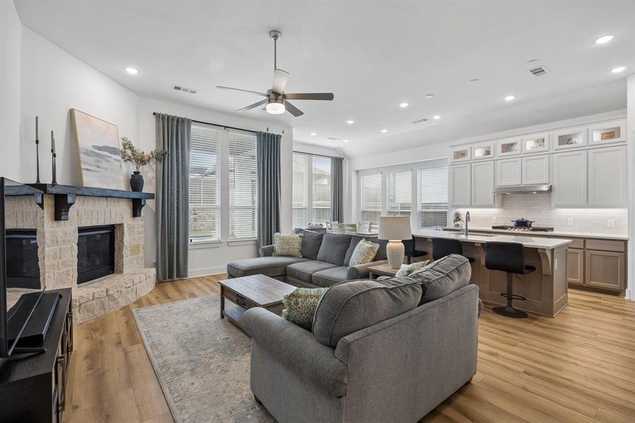 Open-concept living area featuring wood-finish flooring, a stone-clad fireplace with a dark wood mantle, and recessed lighting