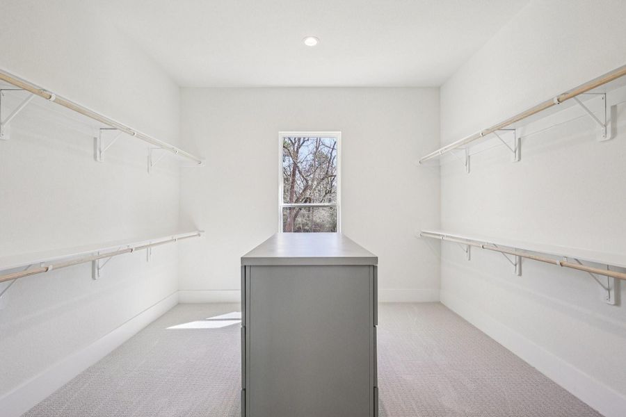 This is a spacious walk-in closet with ample natural light. It features dual rows of shelves on both sides and a central island for additional storage. The neutral color palette creates a bright and airy atmosphere.