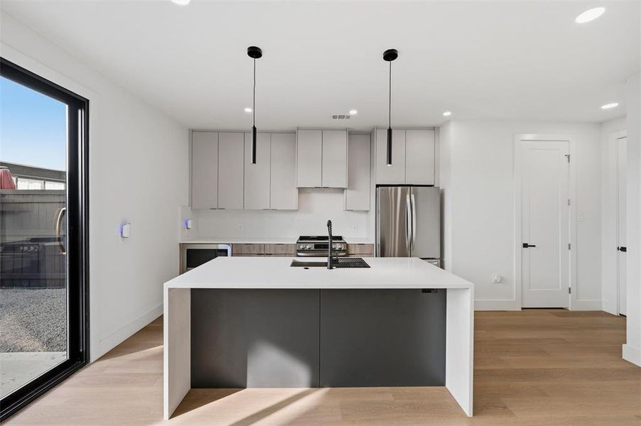 Kitchen features pendant lighting above the island. Light cabinetry provides ample storage throughout the space. Quartz waterfall island includes an undermount sink and black faucet. Stainless steel refrigerator completes the kitchen. Vinyl flooring extends throughout.
