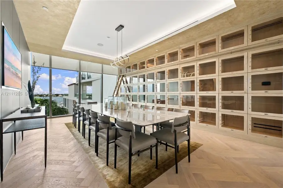 Floor-to-ceiling built-in cabinetry with integrated rolling library ladder, wood-finish herringbone flooring, recessed tray ceiling, modern linear pendant lighting, and full-height glass windows Floor-to-ceiling built-in cabinetry with integrated rolling library ladder, wood-finish herringbone flooring, recessed tray ceiling, modern linear pendant lighting, and full-height glass windows