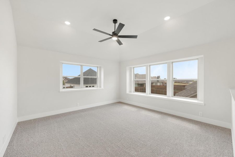 Empty bedroom with gray carpet, white walls, ceiling fan, and multiple windows with neighborhood views