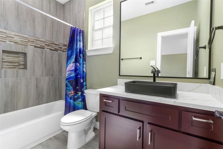 This modern bathroom features a sleek vanity with a black vessel sink and marble countertop. The shower area has stylish tilework and a colorful ocean-themed curtain, while natural light brightens the space through a small window.