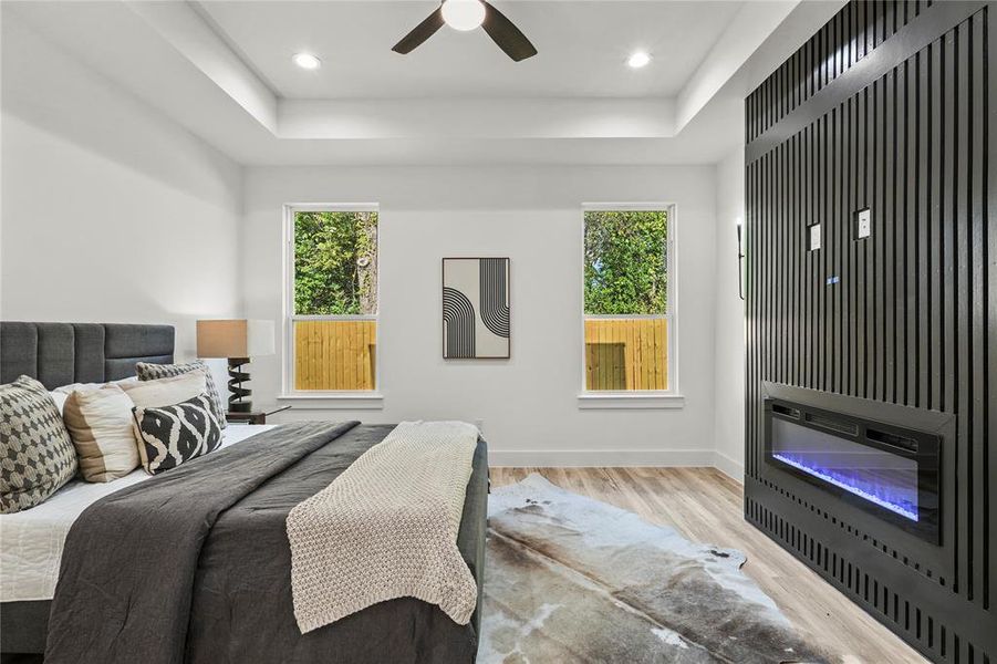 Bedroom featuring a glass covered fireplace, wood finished floors, a tray ceiling, recessed lighting, and a ceiling fan