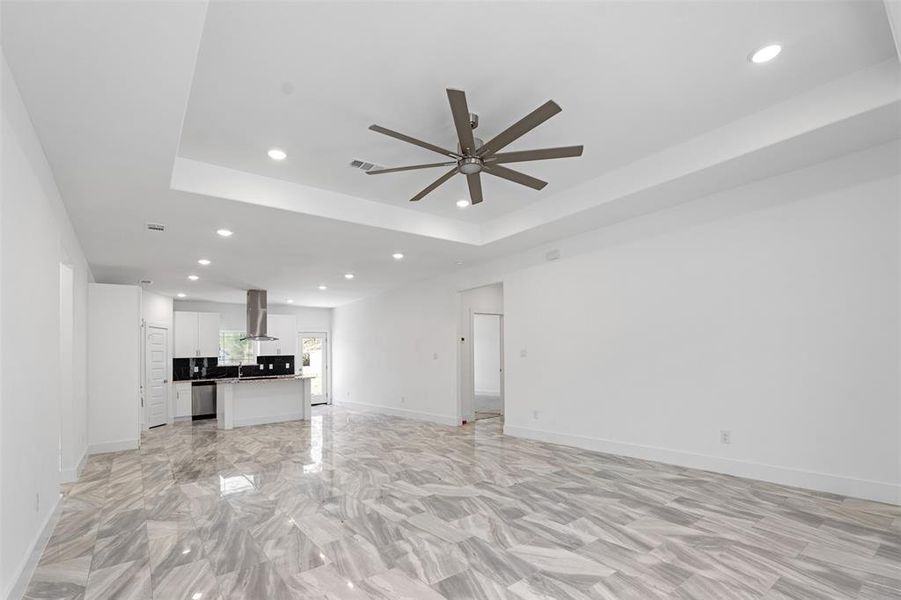 Unfurnished living room featuring a raised ceiling, ceiling fan, and kitchen