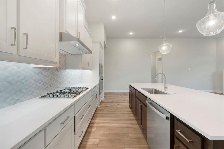 Dual tone kitchen with two tone cabinets, stainless steel appliances, light wood finished floors, hanging light fixtures, and a kitchen island with sink
