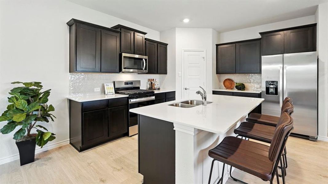 Kitchen featuring appliances with stainless steel finishes, backsplash, light wood finished floors, a center island with sink, and light stone countertops