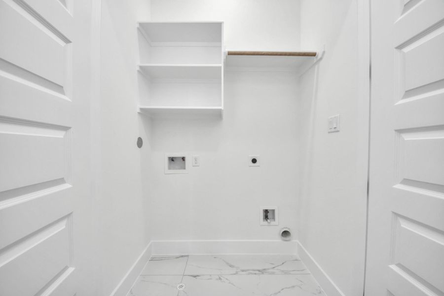 This is a bright, compact laundry room featuring white walls, built-in shelves, hookups for a washer and dryer, and a marble-patterned tile floor.
