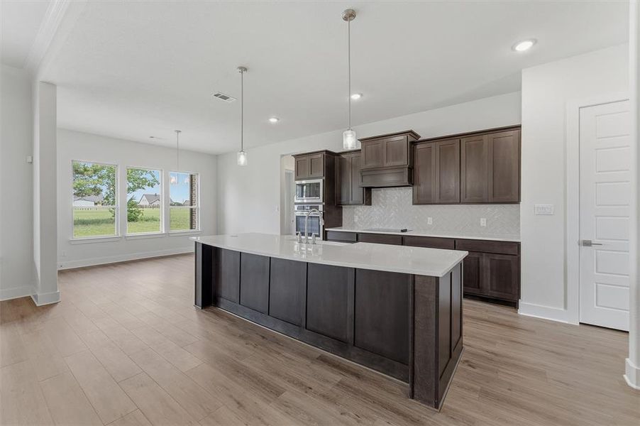 Open-concept kitchen and living area featuring wood-finish flooring, a large kitchen island with a light-toned countertop, dark wood cabinetry, and a white herringbone-patterned backsplash