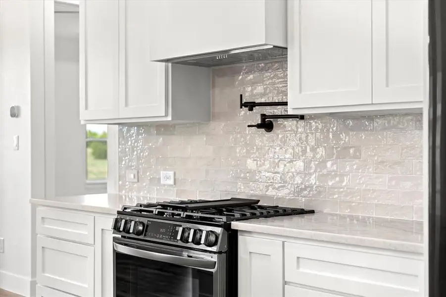 Kitchen with tasteful backsplash, gas stove, wall chimney exhaust hood, and white cabinetry Kitchen with tasteful backsplash, gas stove, wall chimney exhaust hood, and white cabinetry