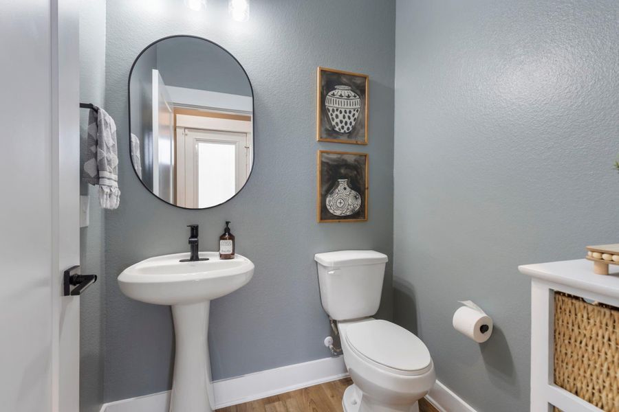 This is a compact, modern bathroom featuring a pedestal sink, toilet, and oval mirror. The walls are painted a soothing gray, complemented by wood flooring and minimalist decor.