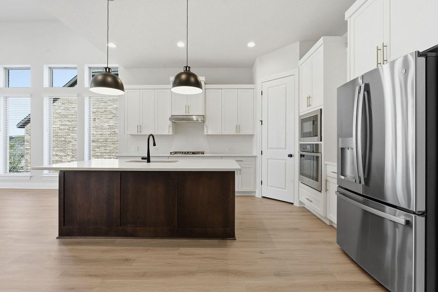 Two tone kitchen featuring stainless steel appliances, decorative light fixtures, light wood finished floors, a kitchen island with sink, and two tone cabinets