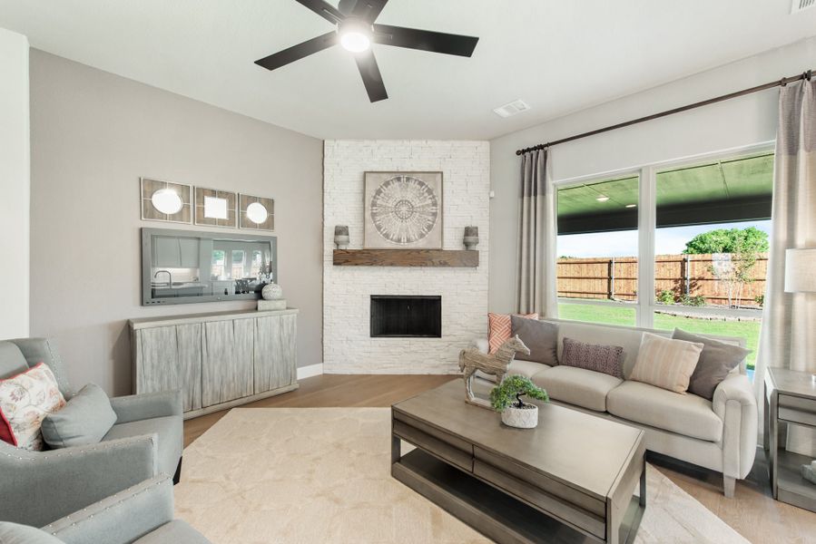Living room with white brick corner fireplace, ceiling fan, gray sofa, and large window overlooking backyard