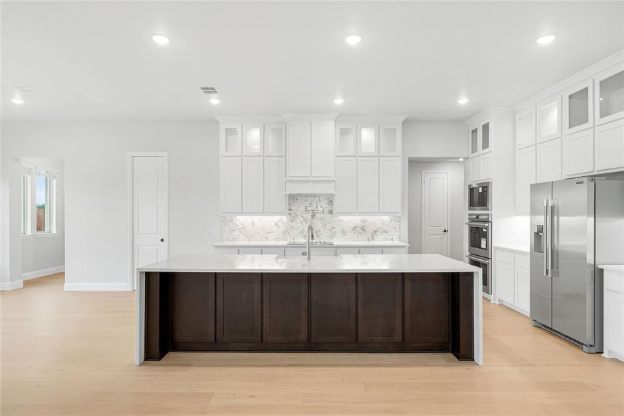 Large kitchen with white cabinets, dark wood island, marble backsplash, and stainless steel appliances on light hardwood floors