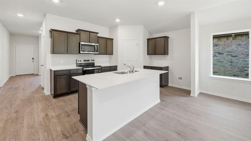 Kitchen with stainless steel appliances, dark brown cabinets, a kitchen island with sink, light wood-type flooring, and recessed lighting Kitchen with stainless steel appliances, dark brown cabinets, a kitchen island with sink, light wood-type flooring, and recessed lighting