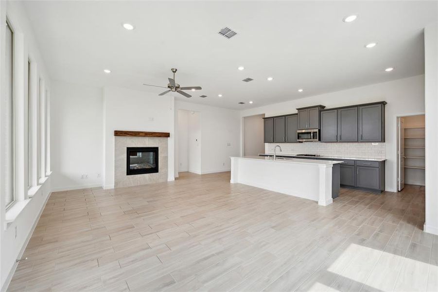 Kitchen featuring a center island with sink, gray cabinetry, a tiled fireplace, ceiling fan, and decorative backsplash