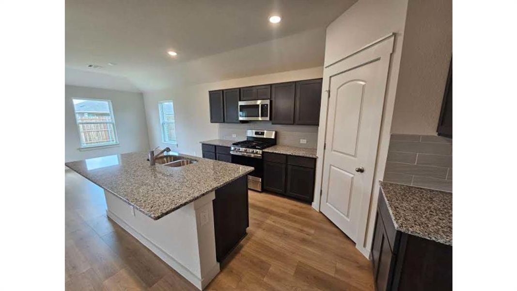 Kitchen featuring a central island with an undermount sink, granite countertops, wood-finish flooring, dark cabinetry, and stainless steel appliances Kitchen featuring a central island with an undermount sink, granite countertops, wood-finish flooring, dark cabinetry, and stainless steel appliances