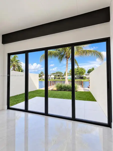 Floor-to-ceiling multi-panel sliding glass doors with matte black framing, exposed structural ceiling beam, polished stone-finish flooring, paved exterior patio, and water frontage Floor-to-ceiling multi-panel sliding glass doors with matte black framing, exposed structural ceiling beam, polished stone-finish flooring, paved exterior patio, and water frontage