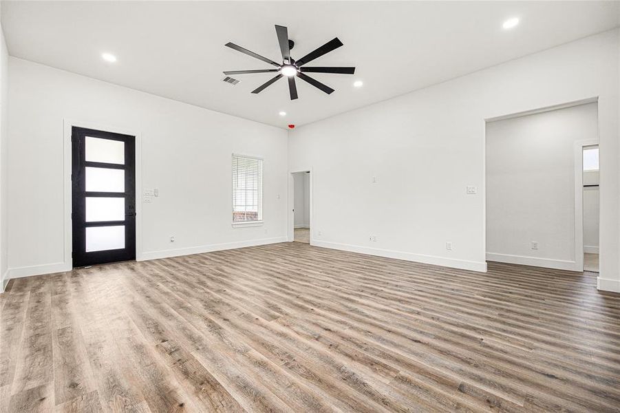 This spacious living area features sleek wood flooring, a modern ceiling fan, and ample natural light from large windows. The room is accented by a stylish black-framed door and clean white walls, creating a bright and inviting atmosphere.