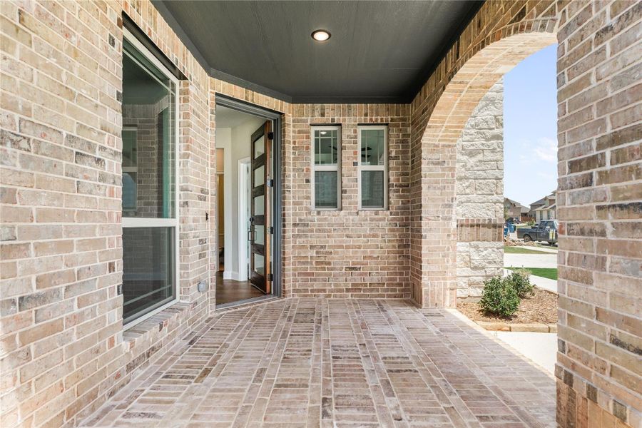 Covered front porch with brick flooring, arched stone column, and open front door with glass panels Covered front porch with brick flooring, arched stone column, and open front door with glass panels