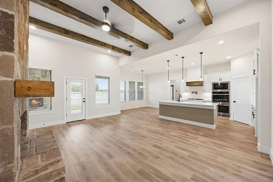 Open-concept living space featuring wood-finish flooring, a stone fireplace with a wood mantle, vaulted ceilings with exposed wood beams, and a contemporary kitchen with a central island and pendant lighting