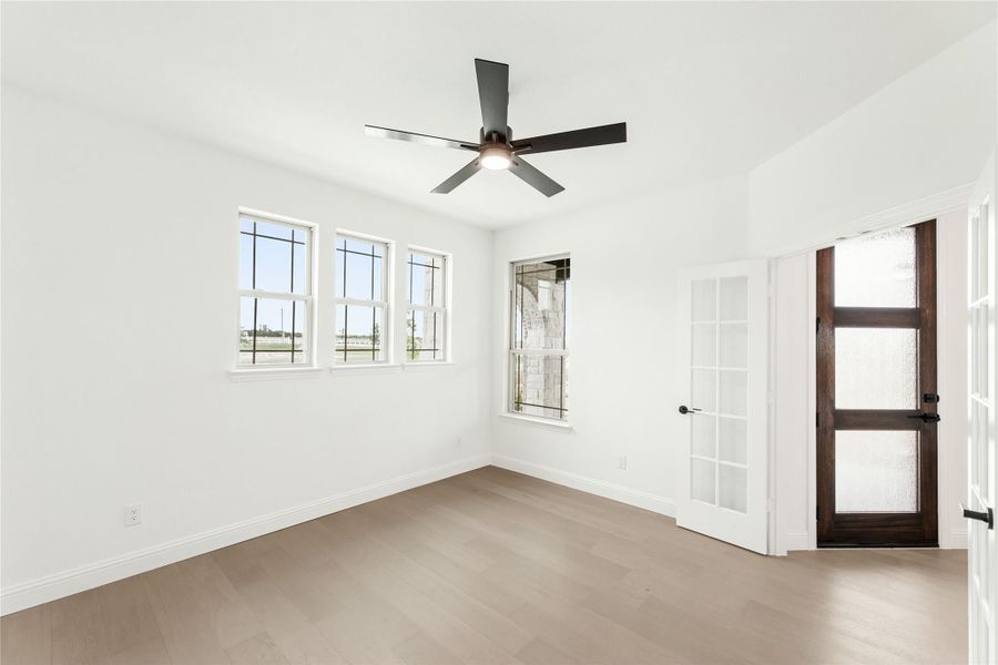 Empty room with light wood floors, ceiling fan, multiple windows, and dark wood glass-panel door Empty room with light wood floors, ceiling fan, multiple windows, and dark wood glass-panel door