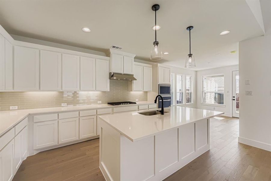 Kitchen featuring pendant lighting, a kitchen island with sink, white cabinetry, light wood-type flooring, and decorative backsplash Kitchen featuring pendant lighting, a kitchen island with sink, white cabinetry, light wood-type flooring, and decorative backsplash