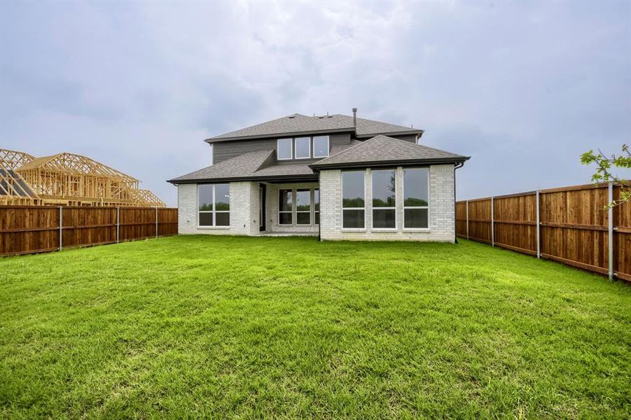 Expansive fenced backyard featuring a verdant lawn, a white brick exterior, a dark-toned siding accent, multiple large windows, and a multi-level roofline Expansive fenced backyard featuring a verdant lawn, a white brick exterior, a dark-toned siding accent, multiple large windows, and a multi-level roofline