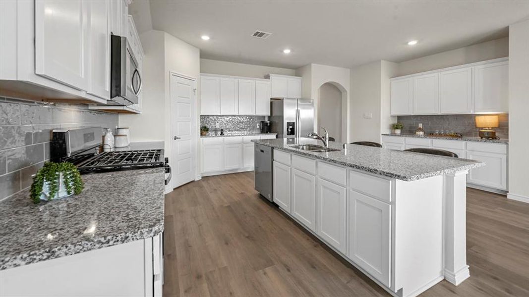 Kitchen featuring white cabinetry, backsplash, stainless steel appliances, light stone countertops, and an island with sink