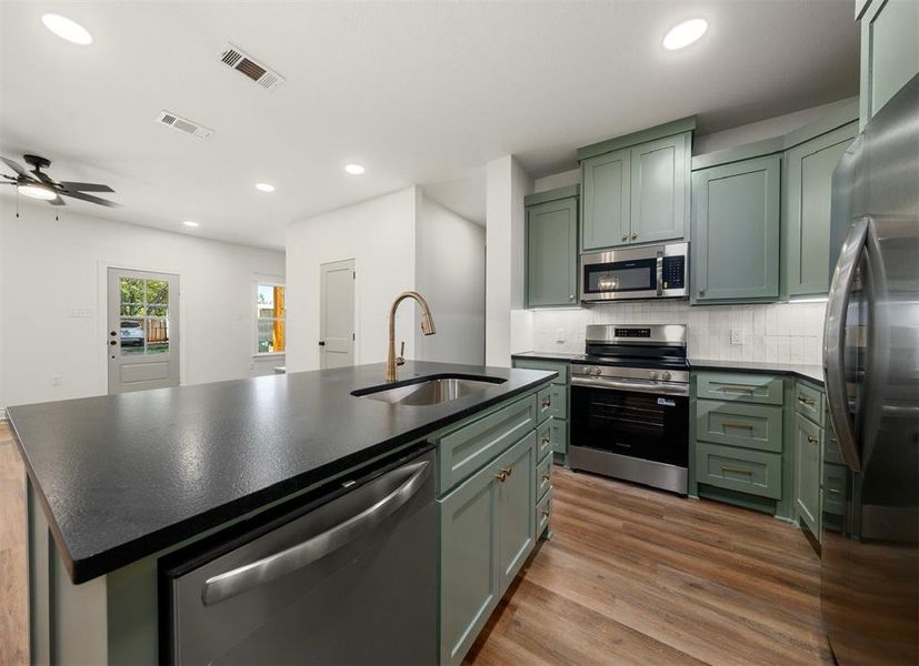 Kitchen featuring green cabinets, appliances with stainless steel finishes, backsplash, light wood-style floors, and a kitchen island with sink Kitchen featuring green cabinets, appliances with stainless steel finishes, backsplash, light wood-style floors, and a kitchen island with sink