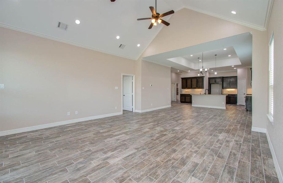 This is a spacious and modern open-plan living area with high ceilings, contemporary wood-look tile flooring, and a view into the kitchen with a large island and dark wood cabinets.