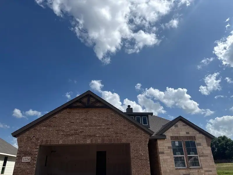 New construction exterior featuring brick and stone cladding, a prominent gable with decorative wood accents, dark shingle roofing, and an integrated two-car garage New construction exterior featuring brick and stone cladding, a prominent gable with decorative wood accents, dark shingle roofing, and an integrated two-car garage