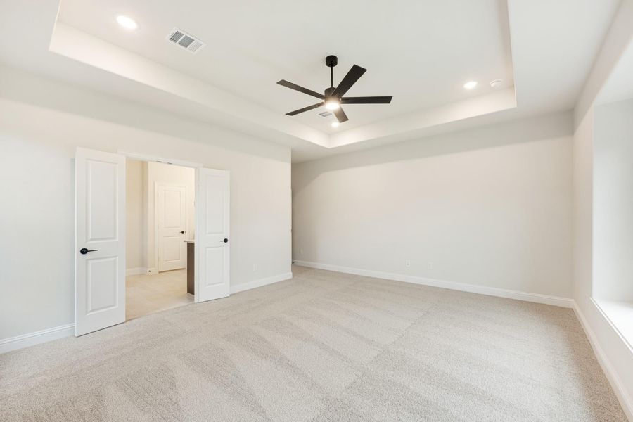 Empty bedroom with tray ceiling, black ceiling fan, carpet flooring, and open door to ensuite bathroom Empty bedroom with tray ceiling, black ceiling fan, carpet flooring, and open door to ensuite bathroom