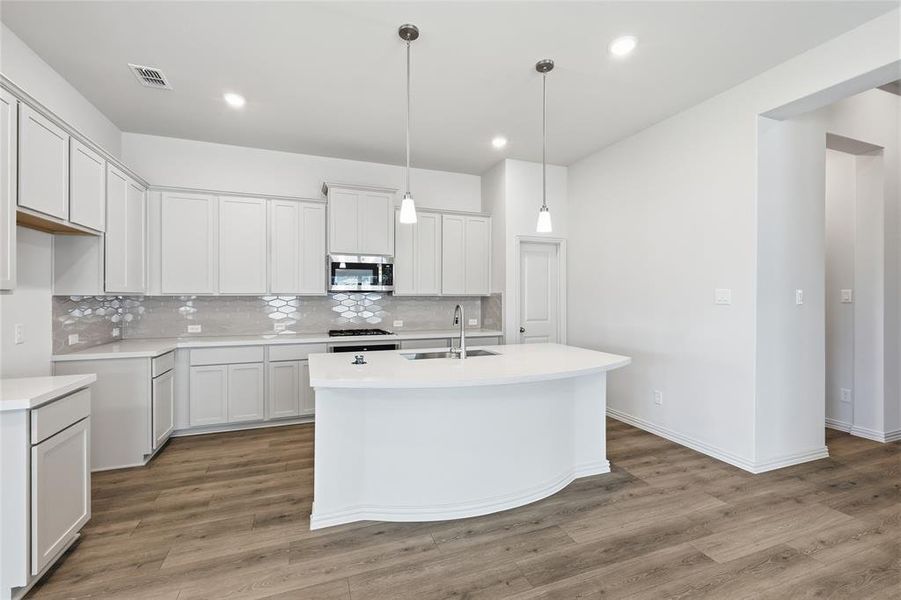 Kitchen featuring light wood-style floors, stainless steel microwave, a kitchen island with sink, and decorative light fixtures