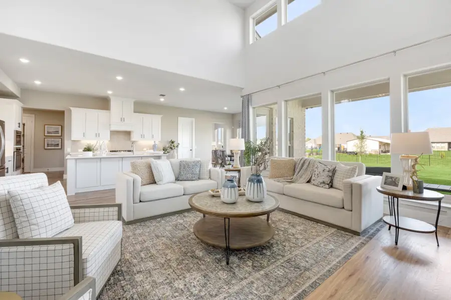 Open-concept living room with high ceilings, large windows, hardwood floors, and white kitchen visible in background. Open-concept living room with high ceilings, large windows, hardwood floors, and white kitchen visible in background.