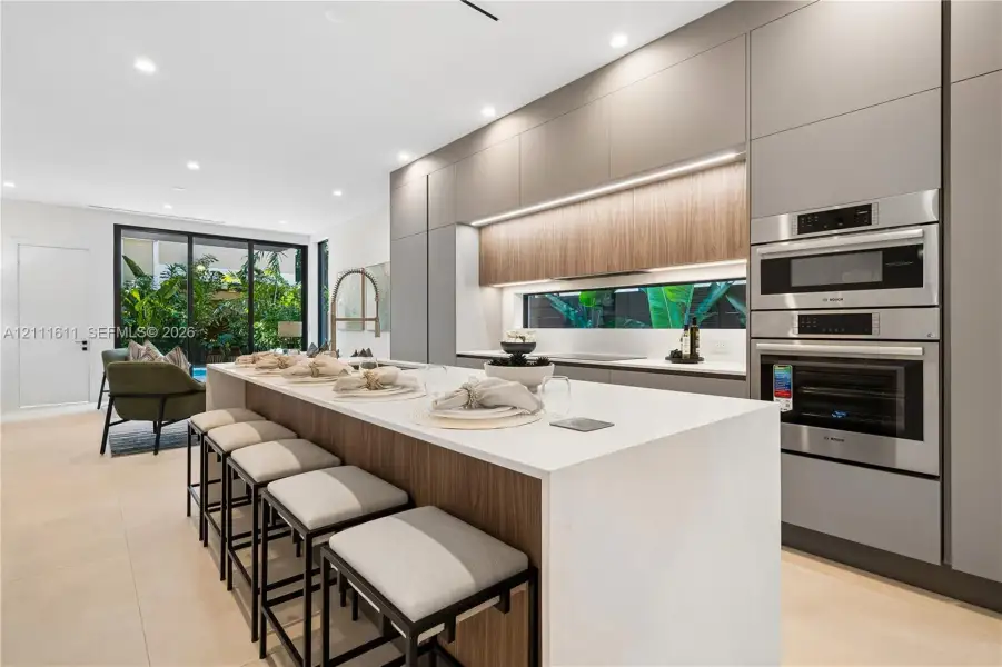 Modern kitchen featuring flat-panel cabinetry, a central island with a solid-surface countertop, integrated stainless steel double wall ovens, recessed lighting, and large-format flooring Modern kitchen featuring flat-panel cabinetry, a central island with a solid-surface countertop, integrated stainless steel double wall ovens, recessed lighting, and large-format flooring