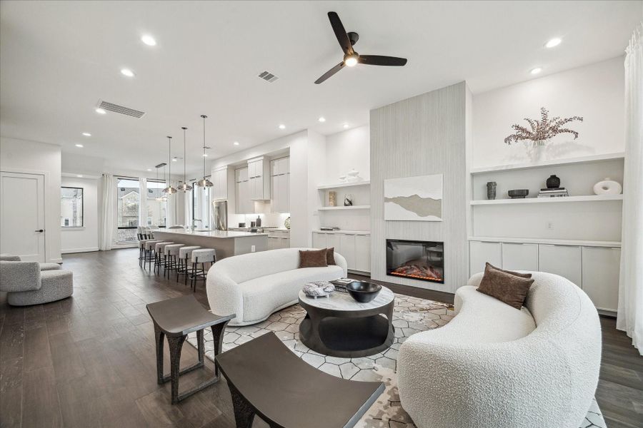 Open-concept second-floor living designed for elevated entertaining, featuring soaring ceilings, wide-plank engineered wood flooring, recessed lighting, and a sleek gas fireplace with custom built-ins. The seamless flow connects living, dining, and kitchen spaces, while oversized windows and balcony access bring in natural light. A designer dry bar with beverage fridge enhances hosting, blending modern elegance with everyday comfort.