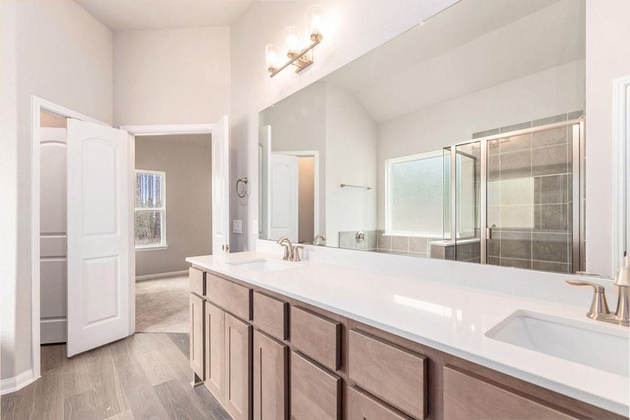 This bathroom features a spacious double vanity with a large mirror, modern light fixtures, and ample storage. It includes a glass-enclosed shower with stylish tile and a frosted window for privacy, offering a bright and inviting space.
