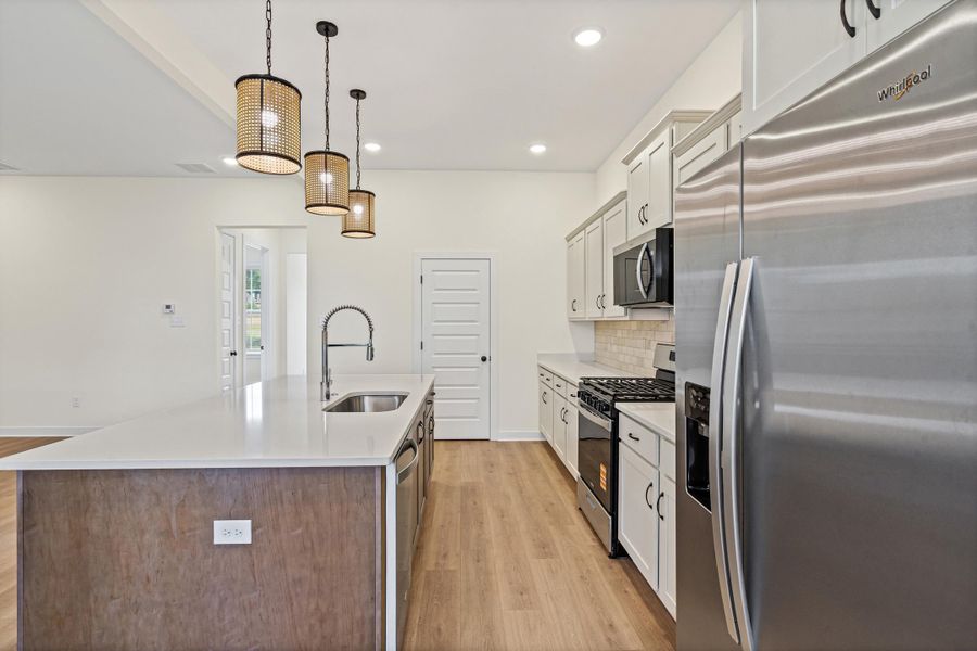Kitchen featuring a spacious island with an undermount sink, pendant lighting, stainless steel appliances, light wood-finish flooring, and white cabinetry Kitchen featuring a spacious island with an undermount sink, pendant lighting, stainless steel appliances, light wood-finish flooring, and white cabinetry