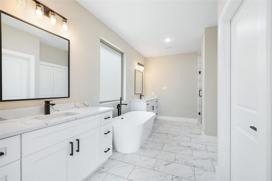 The primary bathroom exudes a sleek, modern design with a luxurious freestanding tub, dual sinks, and elegant marble-inspired flooring. A neutral color palette enhances the bright, inviting atmosphere, while ample lighting highlights the clean lines and sophisticated finishes, creating a serene and relaxing retreat.