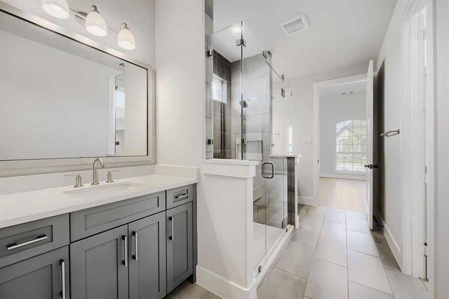 Bathroom featuring a spacious walk-in shower with clear glass enclosure and dark tile surround, a single vanity with grey cabinetry and a light-toned countertop, a rectangular undermount sink with a brushed nickel faucet, and neutral-toned floor tiling