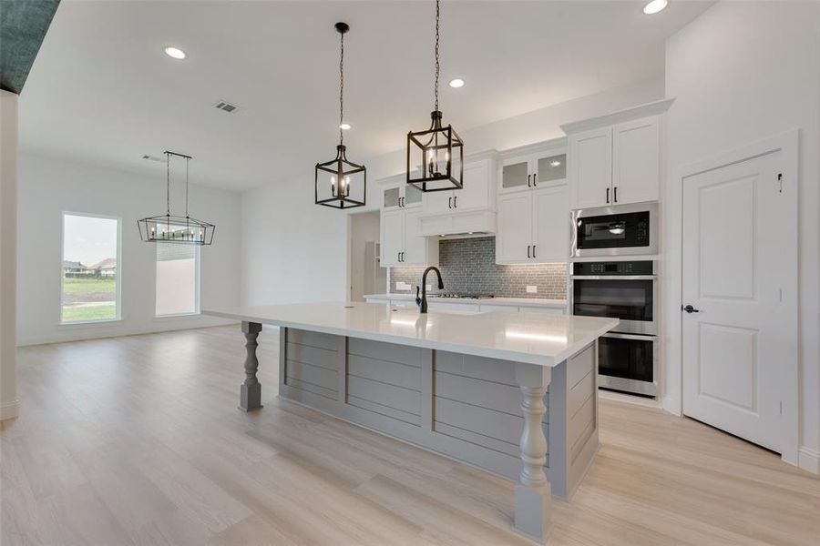 Kitchen featuring stainless steel appliances, hanging light fixtures, decorative backsplash, light wood-type flooring, and an island with sink