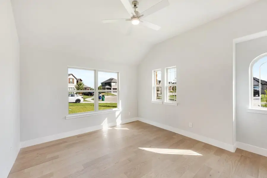 Empty room with vaulted ceiling, ceiling fan, hardwood floors, and large windows overlooking a neighborhood