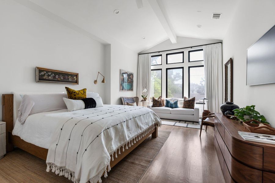 Welcome to the primary suite, tucked away from the main living areas, it features a vaulted ceiling, hardwood flooring throughout, custom drapes, and a stunning wall of oversized windows with upper transom windows.