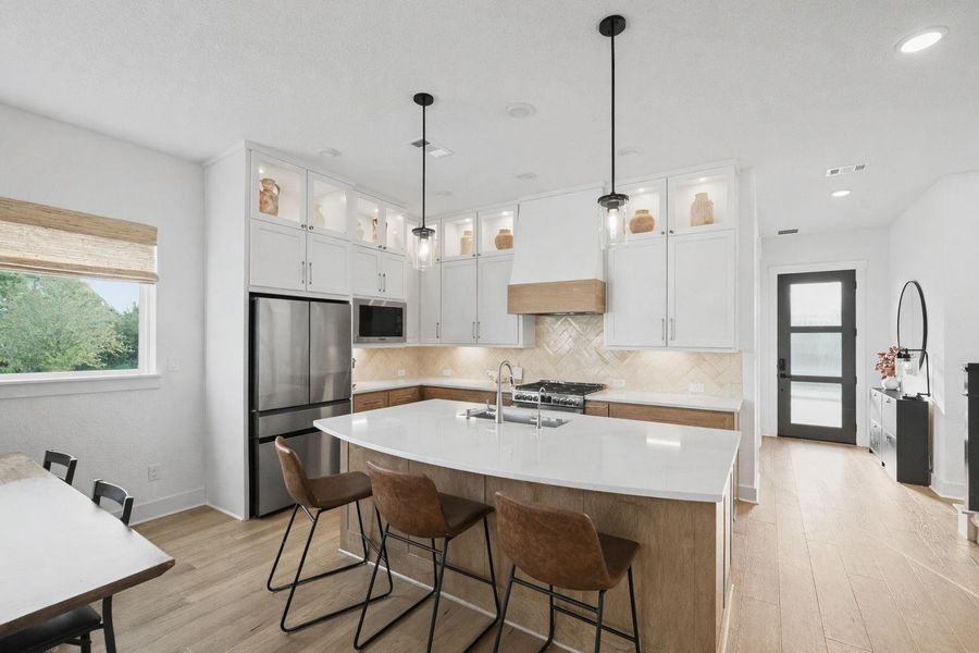 Chef-inspired kitchen showcasing an expansive quartz island with seating, custom cabinetry with illuminated display accents, and sleek stainless appliances. Designer pendant lighting, warm wood flooring, and a stylish herringbone backsplash elevate the space, while the open layout offers a refined blend of luxury and functionality.