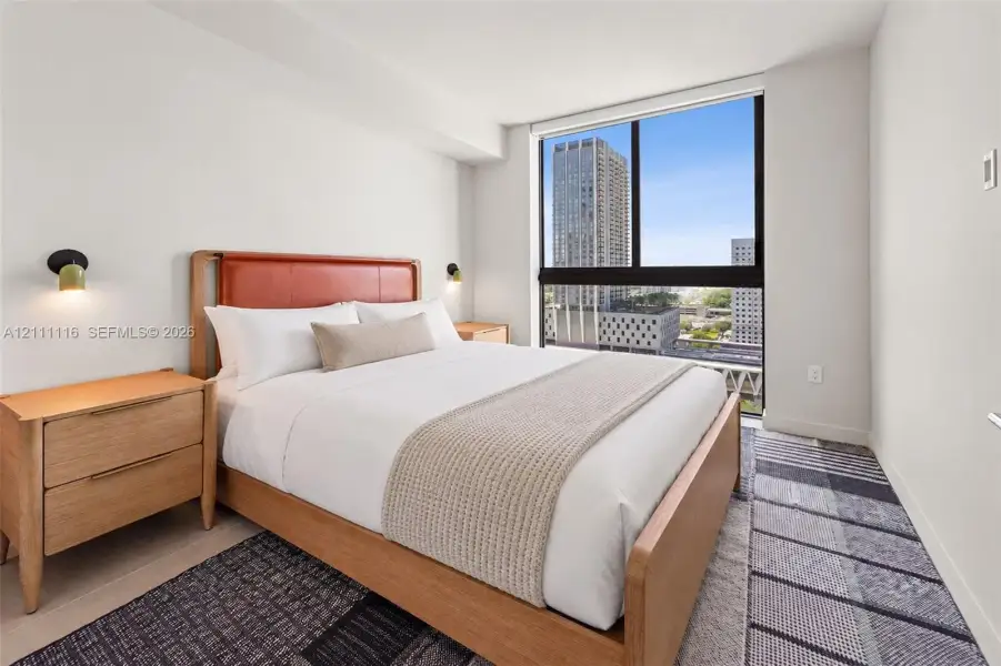 Floor-to-ceiling windows featuring urban scenery, two wall-mounted metallic light fixtures, and neutral interior finishes