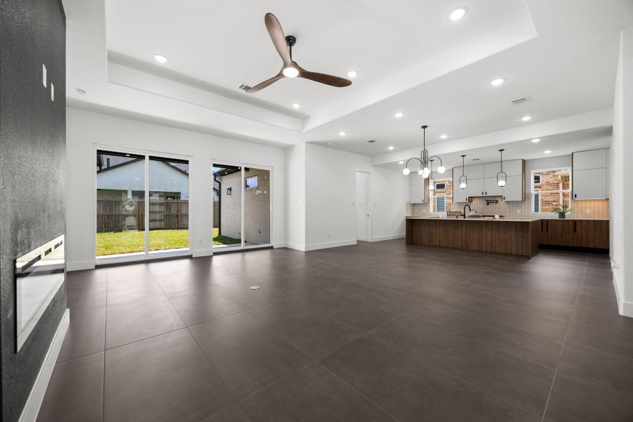 This photo showcases a spacious open-concept living area with modern design elements. It features large tile flooring, recessed lighting, and a ceiling fan. The room seamlessly connects to a stylish kitchen with a large island, contemporary pendant lights, and ample natural light from sliding glass doors leading to a fenced backyard.