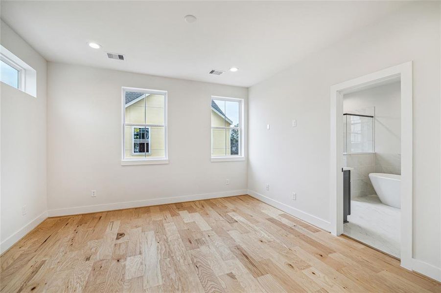 This bright room features light hardwood floors, fresh white walls, and recessed lighting. Large windows allow natural light, and there's an en-suite bathroom with a modern freestanding tub.