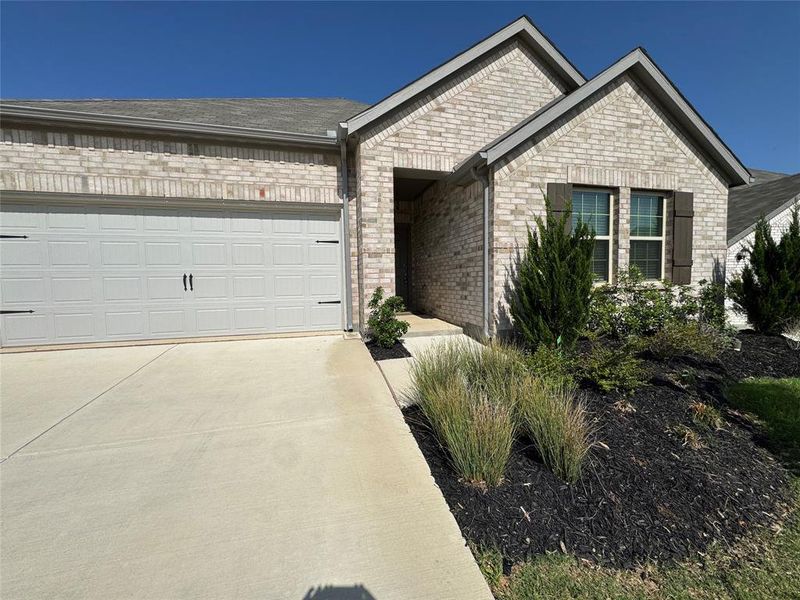 Light-colored brick exterior with a two-car garage, dark decorative window shutters, and low-maintenance landscaping with dark mulch Light-colored brick exterior with a two-car garage, dark decorative window shutters, and low-maintenance landscaping with dark mulch